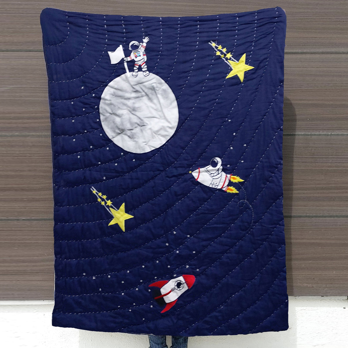 Full view of navy blue quilt with stitched appliqué designs of moon, rockets, astronaut, and stars; perfect for boys’ space-themed bedroom, toddler bedspread, or galaxy nursery bedding.